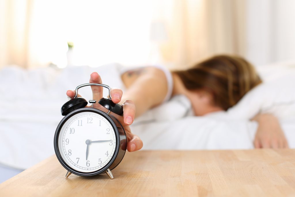 Sleepy young woman trying kill alarm clock while bury face in pillow. Early wake up not getting enough sleep getting work concept. Female stretching hand to ringing alarm willing turn it off