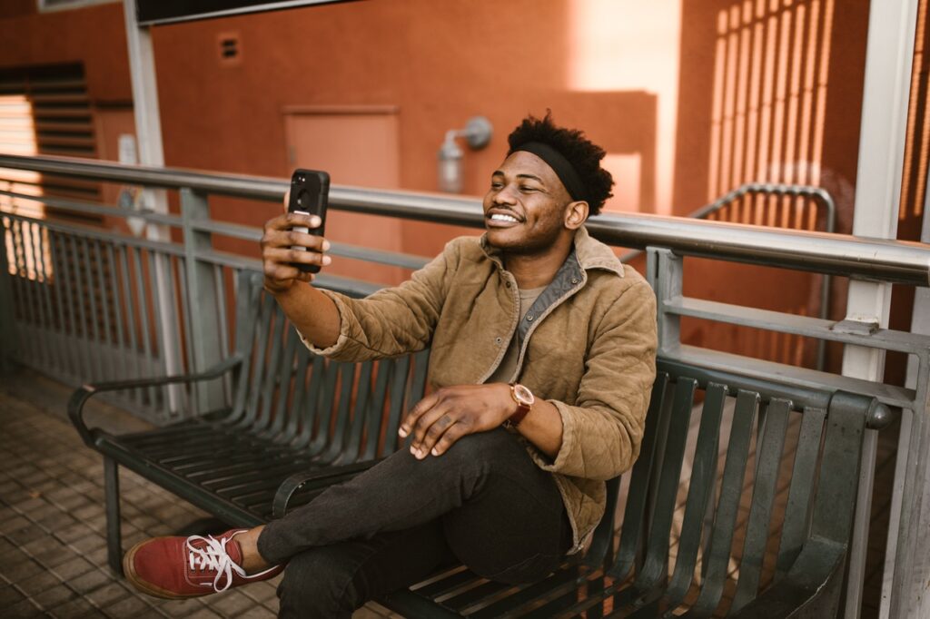 a smiling man sits on a bench holding his phone for a long distance video call or taking a selfie