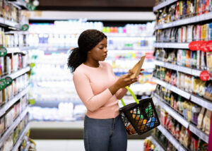 A diverse woman stands in a grocery store aisle holding a basket and looking at a product from the shelf
