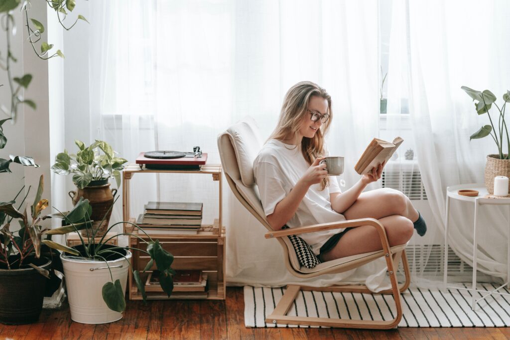 Young woman reading book and sipping coffee while sitting in a chair