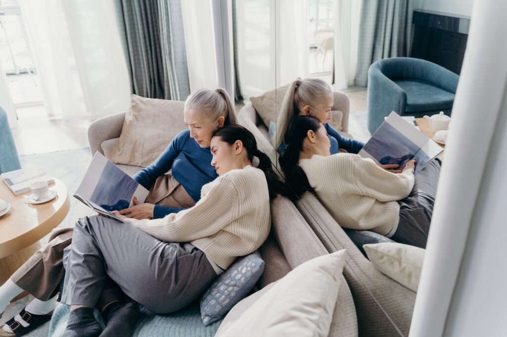 Mother and adult daughter read book together on couch.