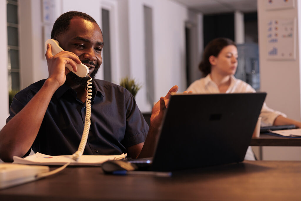 Corporate worker discussing project plan on landline phone, professional office manager answering call, talking with colleague. Smiling man having telephone conversation in coworking space at night