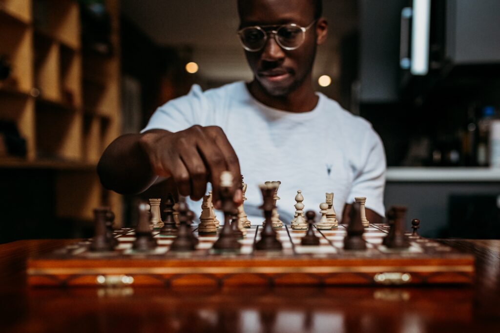 A Black man in a white shirt is playing chess. He is about to move a pawn.