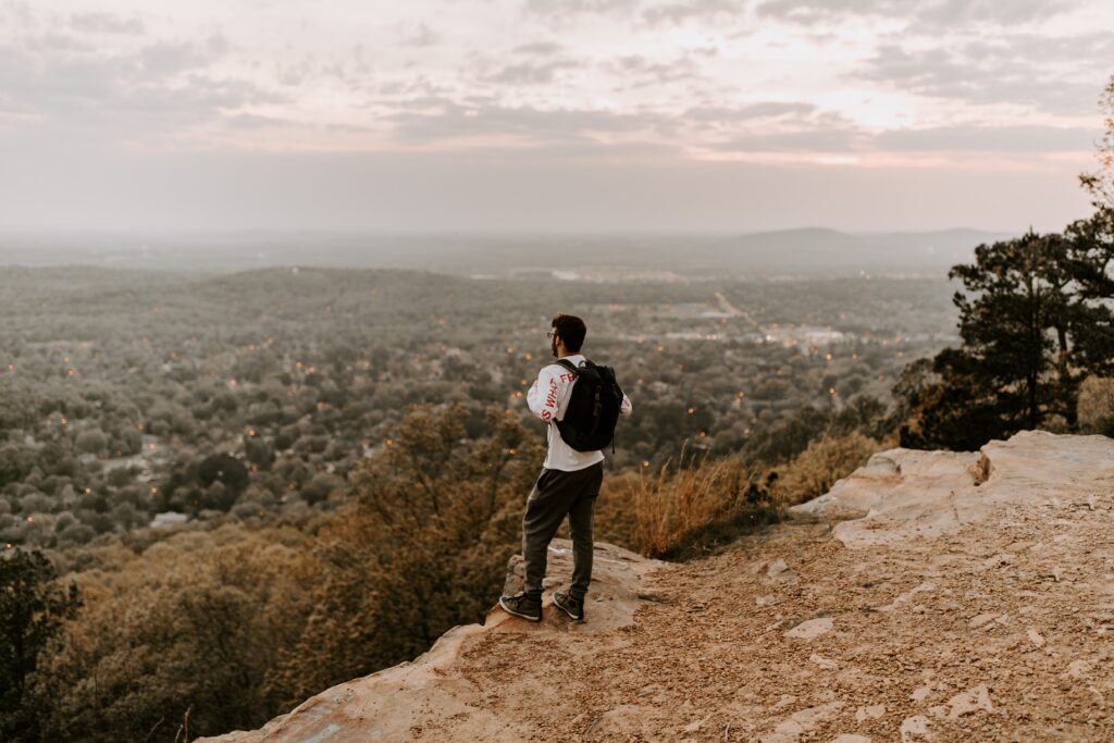 Man standing on the edge of a cliff at sunset looking over a city.