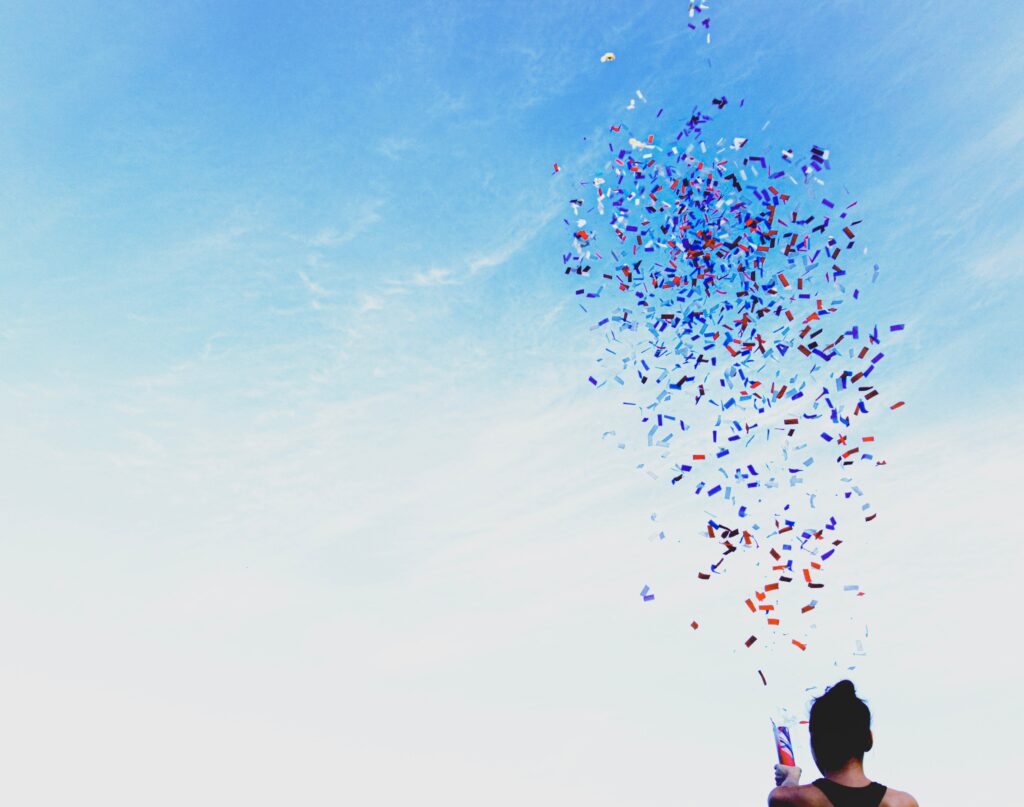 Woman with dark hair in a black tank top located at the bottom right of the screen is popping a confetti popper spraying confetti into the blue sky.