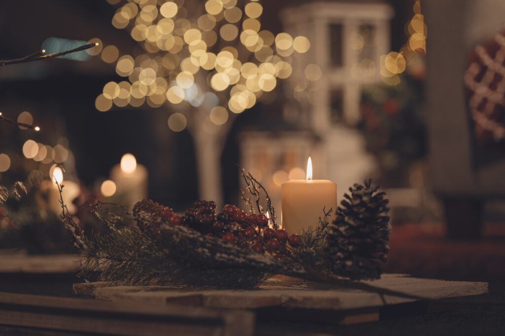 An image of a lit candle on a table surrounded by pine cones in a decorated fashion, with soft lighting from a tree out of focus in the background.