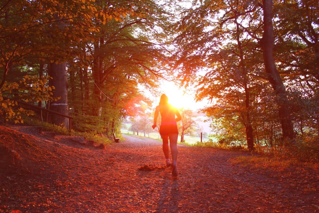 Woman running on a scenic, leaf-covered road in the autumn with the sun on the horizon